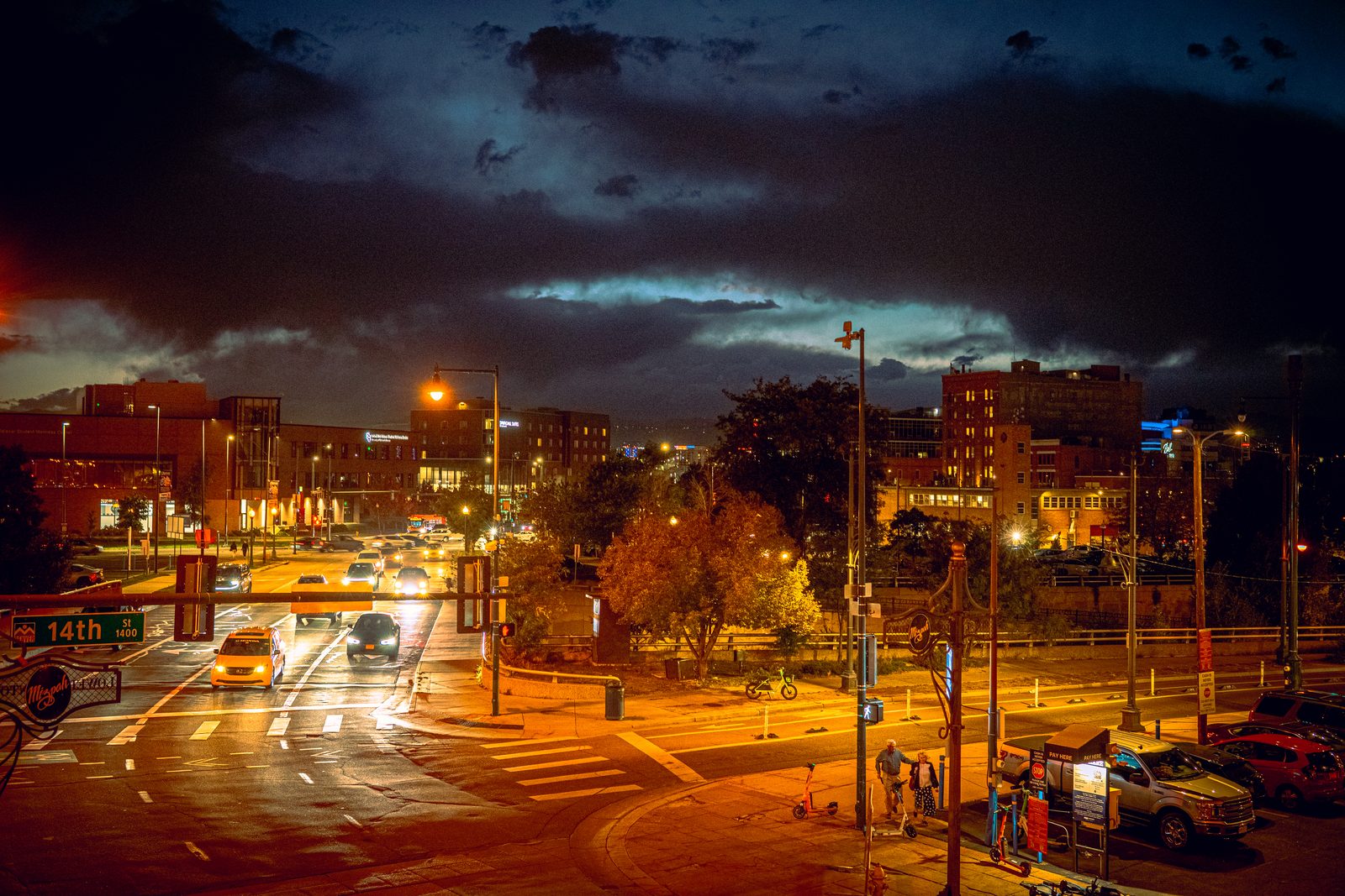 A city street at dusk, light pooling under the traffic signals, a band of orange sky over the rooftops.