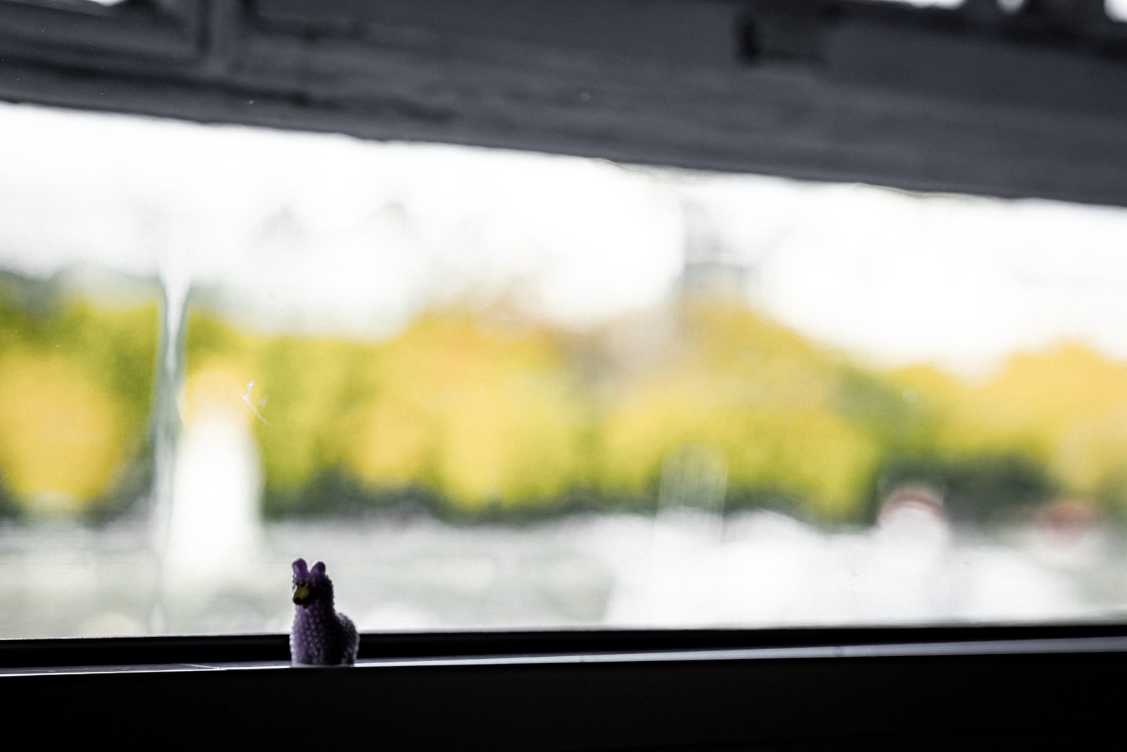 The plastic llama balanced on a dark metal railing with a river and a soft-focus city skyline behind.