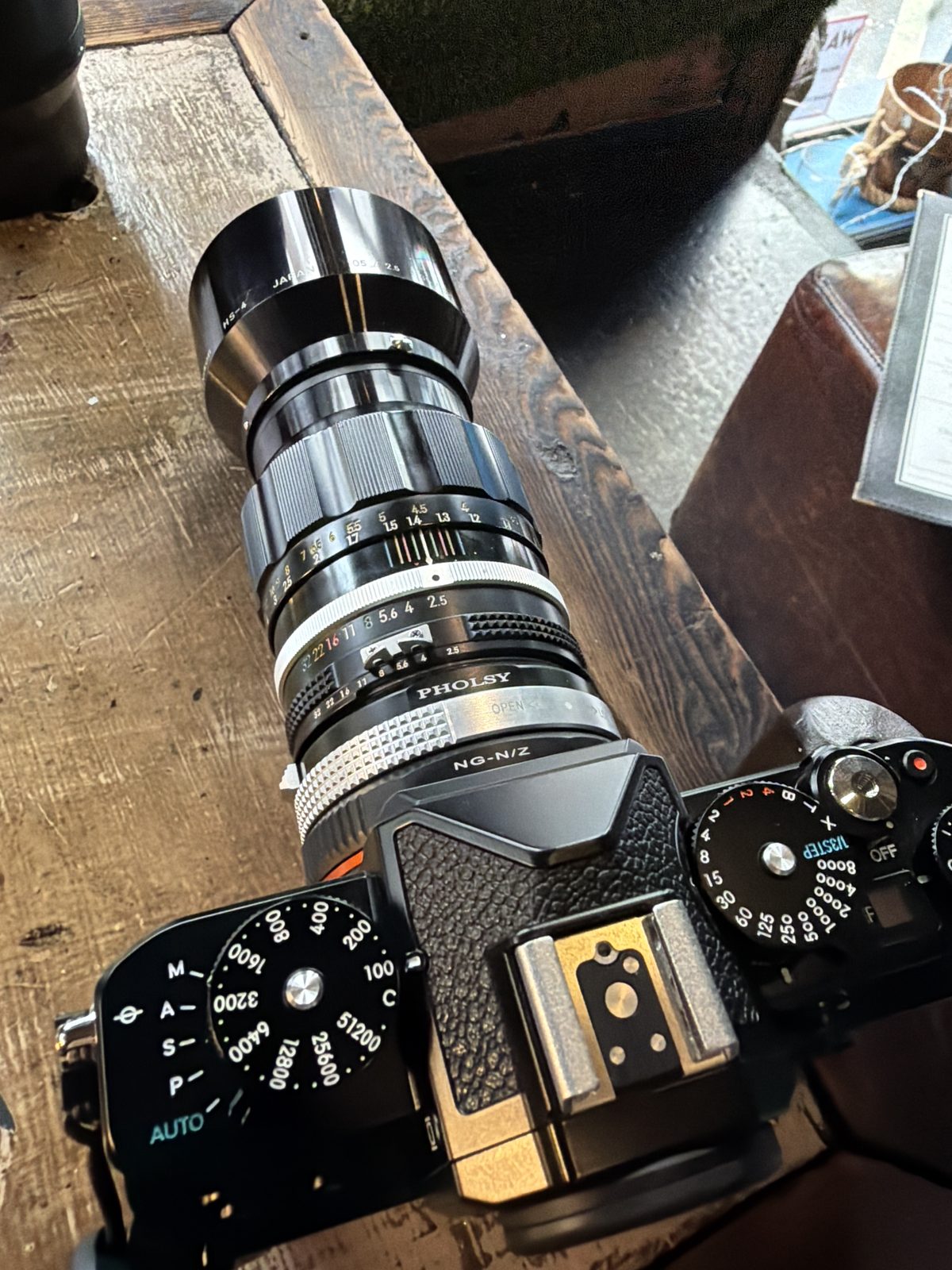 Top-down view of a Nikon Zf with a vintage Nikkor 105mm lens mounted through a Pholsy adapter, on a wood surface, the camera's ISO and shutter dials visible.