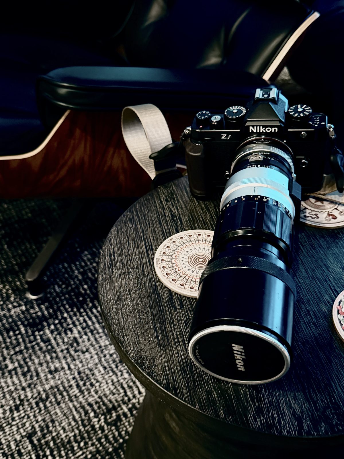 A Nikon Zf fitted with a long vintage 300mm telephoto lens, sitting on a dark round table with a mid-century leather chair behind it.